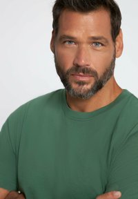 T-shirt verde a maniche corte con scollo rotondo, indossato da un uomo con barba. Tessuto liscio, texture minima e senza motivi visibili.