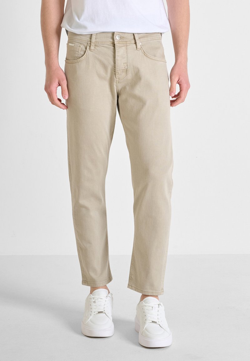 Jeans de algodón beige de pierna recta con diseño de cinco bolsillos y cierre de botón, combinados con zapatillas blancas sobre un fondo neutro.
