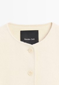 Crème-kleurige wollen jas met ronde halslijn, twee zichtbare knopen en een zwart label met de tekst "Massimo Dutti" aan de binnenkant van de kraag.