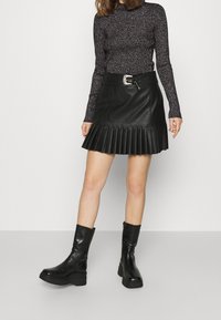 Jupe plissée en cuir noir avec une ceinture ajustée et une boucle argentée, associée à un haut noir texturé à manches longues et des bottines noires.