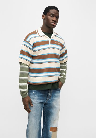 Άνδρας που φορά ριγέ καφέ, μπλε και κρεμ κοντομάνικο πόλο πάνω από χακί μακρυμάνικο πουκάμισο, σε συνδυασμό με μπλε τζιν με φθορές και μπαλώματα.