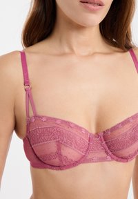 Soutien-gorge en dentelle rose avec tissu transparent, armatures et bretelles réglables. Le design comprend des motifs de dentelle complexes et un léger éclat.