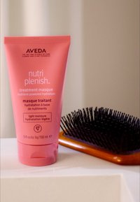 Aveda Nutri Plenish behandlingsmask i en rosa tub, 150 ml. Åtföljd av en hårborste med svarta borststrån på en träbotten.