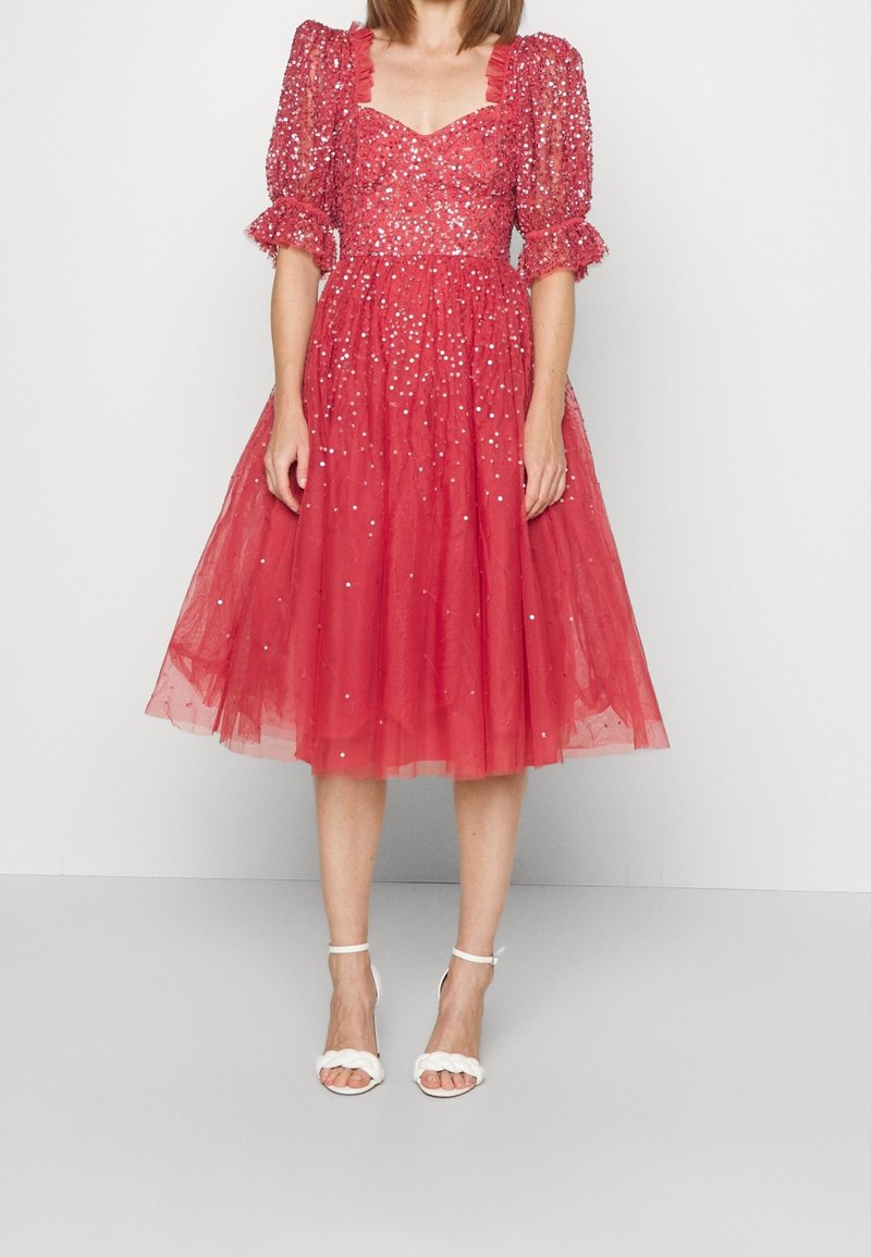 Femme portant une robe rouge en tulle longueur genoux avec manches bouffantes et sequins argentés, associée à des sandales blanches tressées avec brides à la cheville.