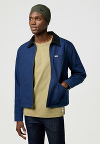 Chaqueta azul marino con cuello negro, cremallera frontal y bolsillos laterales; combinada con una camisa verde oliva. Modelo lleva un gorro oliva.