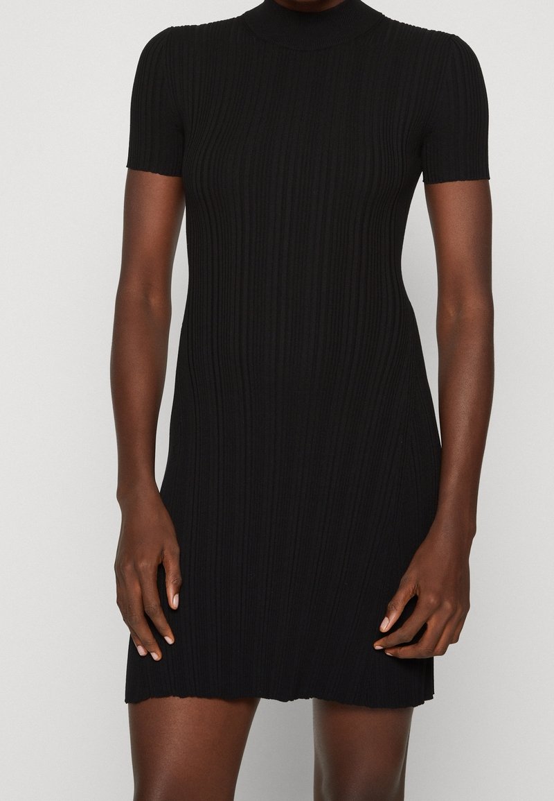 Robe courte ajustée à manches courtes en maille côtelée noire portée par une personne, avec un col haut et un ourlet droit au-dessus des genoux.