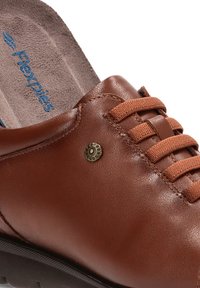Zapato de cuero marrón con punta redondeada, lengüeta texturizada, cordones marrones y un acento decorativo de metal en el lateral. Marca Flexpies visible en el interior.