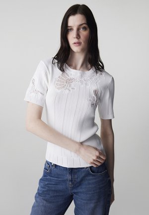 Jeune femme aux cheveux foncés portant un haut tricoté blanc à manches courtes avec des détails en dentelle florale et un jean bleu, posant devant un fond uni.