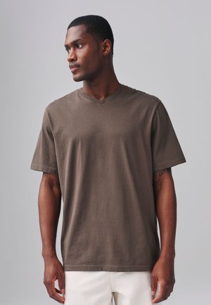 Lühikeste juustega mees, kes kannab pruuni V-kaelusega t-särki ja valgeid pükse, vaadates vasakule ühevärvilise halli tausta ees.