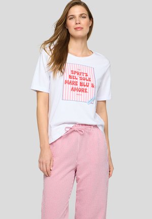 Frau mit langen Haaren, die ein weißes T-Shirt mit rotem Text und rosa gestreifte Hose mit gebundenem Bund vor grauem Hintergrund trägt.