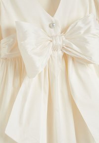 Robe crème avec un large nœud en satin à la taille, une jupe froncée, un décolleté en V, et un accent de bouton en perles. Texture lisse.
