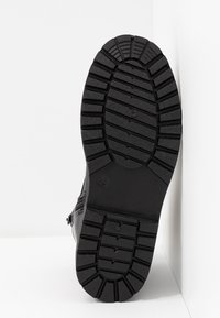 Svart sko med en texturerad gummisula som har djupa slitmönster. Skon visas från undersidan, där storleksmarkeringar syns.