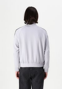 Lichtgrijze zip-up jas met een opstaande kraag, elastische tailleband en zwarte zijstrepen, gemaakt van gladde stof. Heeft lange mouwen en ribbelmanchetten.