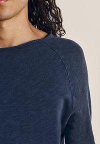 Tamsiai mėlyna ilgomis rankovėmis marškiniai, pagaminti iš minkštos medžiagos, su apvaliu kaklu ir matomomis siūlėmis ant peties siūlės.