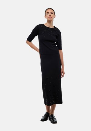 Robe deux pièces noire en tissu tricoté. Manches courtes, haut ajusté avec des embellissements texturés et une longue jupe. Portée avec des chaussures noires.