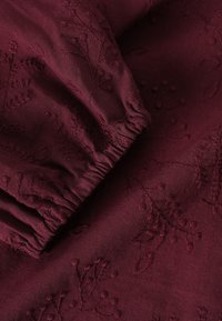 Burgundy kangas dengan pola bunga bordir. Karet yang dijahit di bagian pergelangan tangan. Tekstur lembut dengan finishing halus, menyoroti jahitan yang detail.