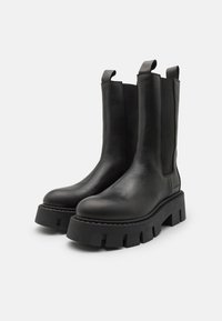 Bottines en cuir noir avec une semelle en caoutchouc texturé et épaisse. Présentent des panneaux latéraux élastiques et des tirettes pour un enfilage facile. Surface lisse.