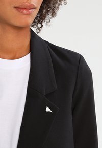 Black blazer with notched lapels, paired with a white T-shirt. A white bird pin is attached to the blazer's lapel, adding detail.