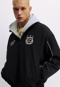 Zwarte jas met ritssluiting en grijze capuchon, met geborduurde logo's en contrasterende witte bies langs de mouwen. Zachte, gestructureerde stof.