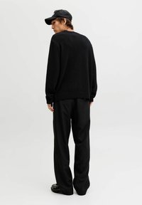 Persona con gorra negra, suéter negro oversize y pantalones negros holgados de rayas, de pie de espaldas sobre un fondo blanco.