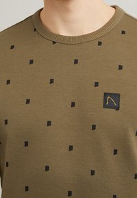 Maglione verde oliva con una maglia strutturata, caratterizzato da motivi a quadrati neri e una piccola toppa nera con un logo arancione sul petto.