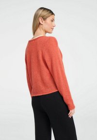 Rūsgans krāsā krāsots īsām piedurknēm džemperis, izgatavots no mīksta, pūkaina auduma, ar brīvu piegriezumu un noapaļotu kakla izgriezumu. Valkāts virs melnām biksēm.