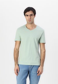 Helles grünes V-Ausschnitt-T-Shirt aus weichem Stoff, mit kurzen Ärmeln und schmaler Passform. Kombiniert mit hellblauen Jeans, vor einem weißen Hintergrund stehend.
