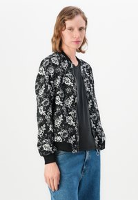 Veste bomber noire avec broderies florales blanches, poignets et ourlet côtelés. Portée sur un t-shirt noir, associée à un jean bleu clair.