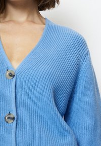 Ljusblå stickad cardigan med V-ringning, vida ärmar och två knappar. Texturen är ribbad med ett mjukt, mysigt utseende.