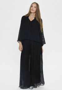 Giovane donna in una lunga blusa nera plissettata e pantaloni a gamba larga che si trova davanti a uno sfondo bianco semplice.