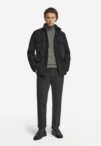 Svart jacka med blixtlås och knappar, två framlommor, bärs över en grå polo, matchad med mörka byxor och svarta skor.