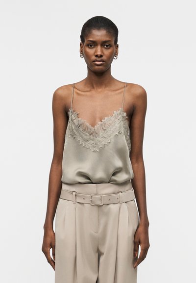 Femme portant un caraco en satin beige avec bordure en dentelle et un pantalon beige taille haute avec ceinture assortie, debout devant un fond uni.