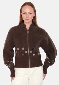 Brun genser med glidelås, med ribbede mansjetter og kant, prydet med et gjentakende logo-mønster i lysere brun farge, laget av et mykt, teksturert materiale.