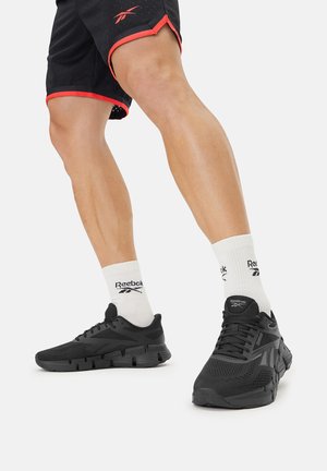 Juodi sportiniai batai su tekstūruotu viršumi, kuriame yra tinklelio dizainas ir paminkštinta padas. Dėvimi su baltais kojinėmis, ant kurių matomas Reebok logotipas.