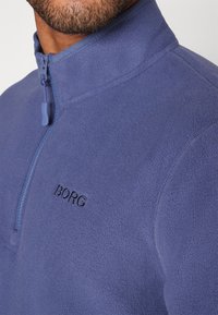 Pullover in pile blu con collo a zip, caratterizzato da cuciture tono su tono e da un logo "BORG" ricamato sul petto. Texture morbida e vestibilità aderente.