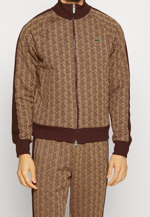 Homme portant une veste zippée marron à motifs et un pantalon assorti avec des bordures marron foncé et de petits logos verts d’alligator sur la poitrine et la cuisse.