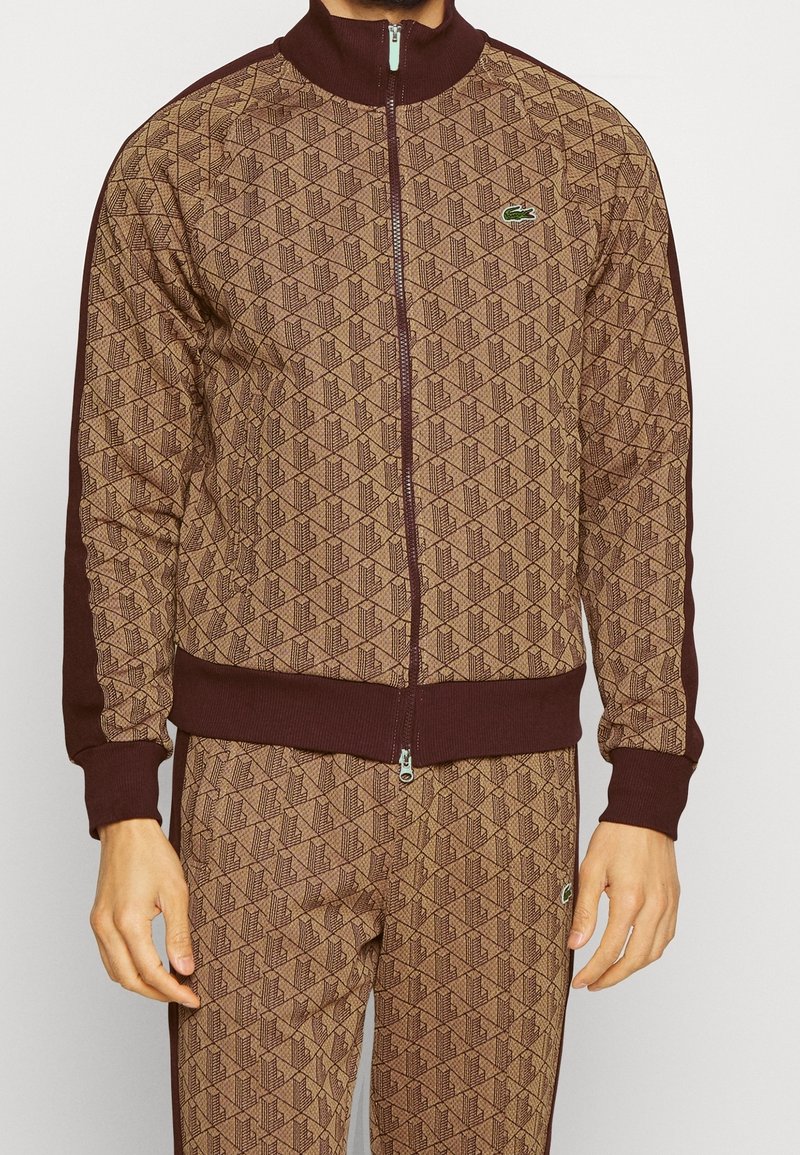 Homme portant une veste zippée marron à motifs et un pantalon assorti avec des bordures marron foncé et de petits logos verts d’alligator sur la poitrine et la cuisse.