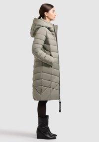 Vaaleanharmaa polvipituinen toppatakki, jossa on huppu ja vinot tikatut kuviot, sekä edessä vetoketju. Yhdistetty mustiin kiilakorkoisiin saappaisiin.