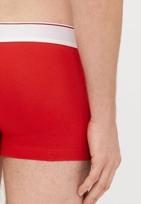 Sarkani bokseršorti ar pieguļošu dizainu. Elastīga josta ar baltu apmali un sarkaniem svītrām. Redzama mīksta, gluda auduma tekstūra.
