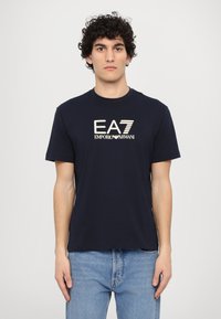 Temno modra majica iz bombaža, z okroglim izrezom, kratkimi rokavi in belim logotipom "EA7 EMPORIO ARMANI" na sprednji strani.