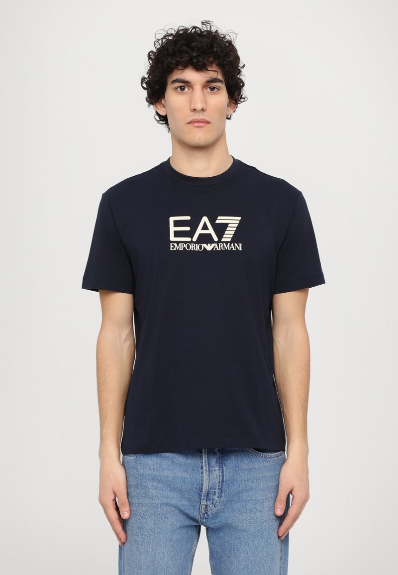 Temno modra majica iz bombaža, z okroglim izrezom, kratkimi rokavi in belim logotipom "EA7 EMPORIO ARMANI" na sprednji strani.
