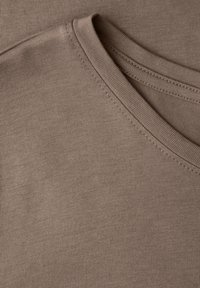 Jasnobrązowa, krótka, materiałowa koszulka o gładkiej fakturze i okrągłym dekolcie, z subtelnymi przeszyciami wzdłuż krawędzi.