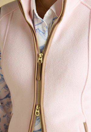 Nahaufnahme einer hellrosa Fleeceweste mit braunem Rand und zwei goldfarbenen Reißverschlüssen, getragen über einem weißen Hemd mit Knöpfen und blauem Vogelmuster.
