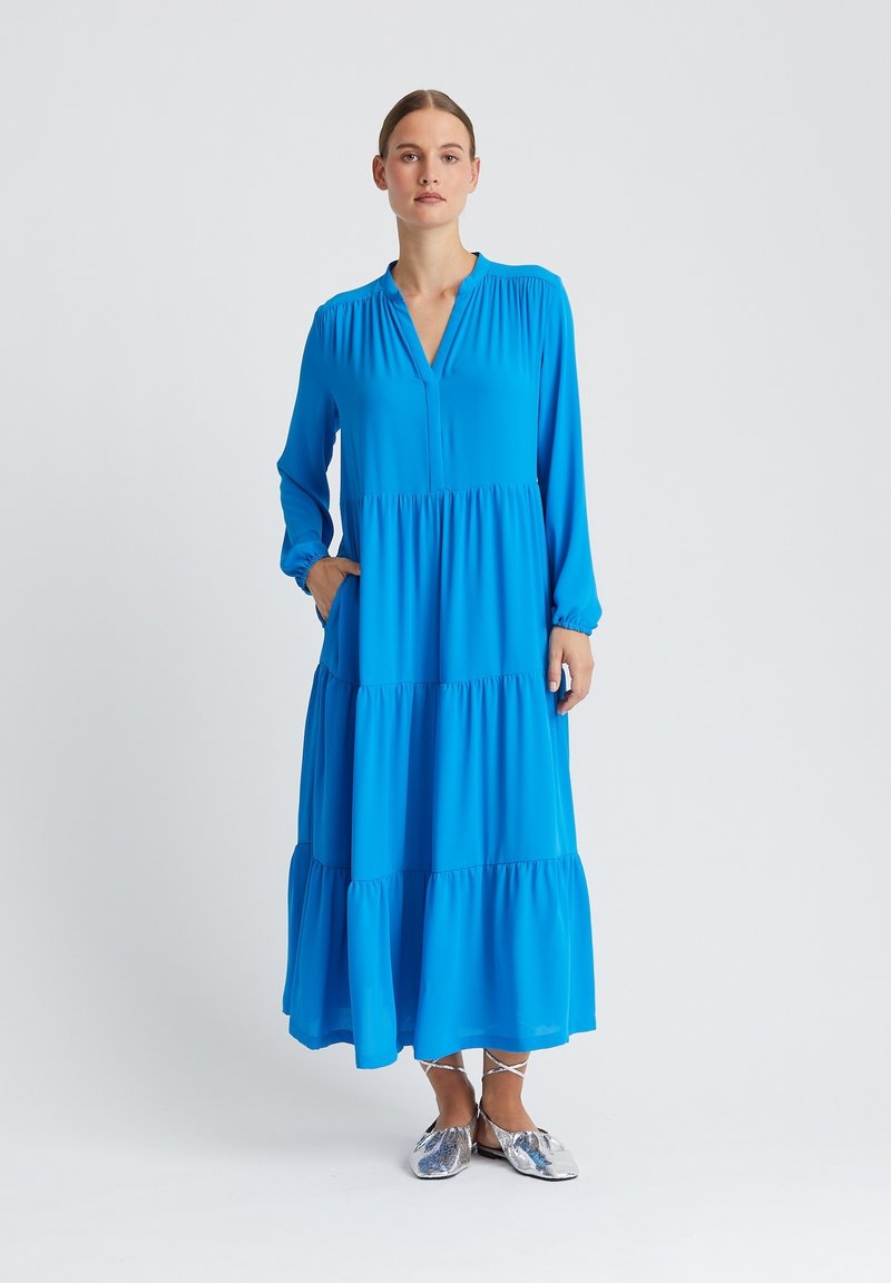Femme portant une robe maxi longue à manches longues, de couleur bleu vif à étages, avec des chaussures plates argentées, debout devant un fond uni clair.