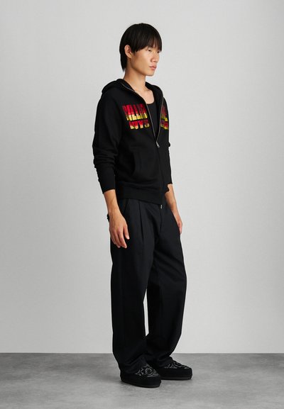 Crna zip-up hoodie s raznobojnim otisnutim tekstom, u paru s labavim crnim hlačama i crnim cipelama s cvjetnim uzorkom.