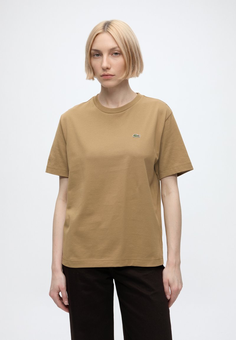 Jeune femme aux cheveux courts et blonds portant un t-shirt beige à col rond et un pantalon noir, debout devant un fond clair uni.