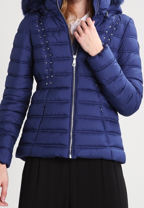 Tmavě modrá prošívaná bunda na zip vpředu, s vertikálním prošitím, zdobená kovovými cvočky a kapucí lemovanou umělou kožešinou.