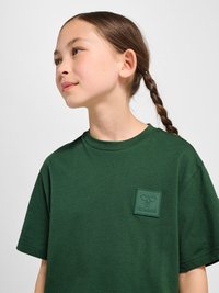 Jeune fille aux cheveux tressés portant un t-shirt vert foncé avec un écusson logo sur la poitrine, regardant vers la gauche sur un fond uni.
