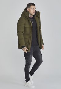 Olivengrøn pufferjakke med hætte, lynlås og forlommer, båret over en sort ribbet sweater og sorte cargo-bukser med hvide sneakers.