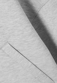 Εγγύς πλάνο μιας τσέπης από ανοιχτό γκρι ύφασμα με ορατή ραφή και πολλαπλές στρώσεις, που αναδεικνύει την υφή και τις λεπτομέρειες του ενδύματος.